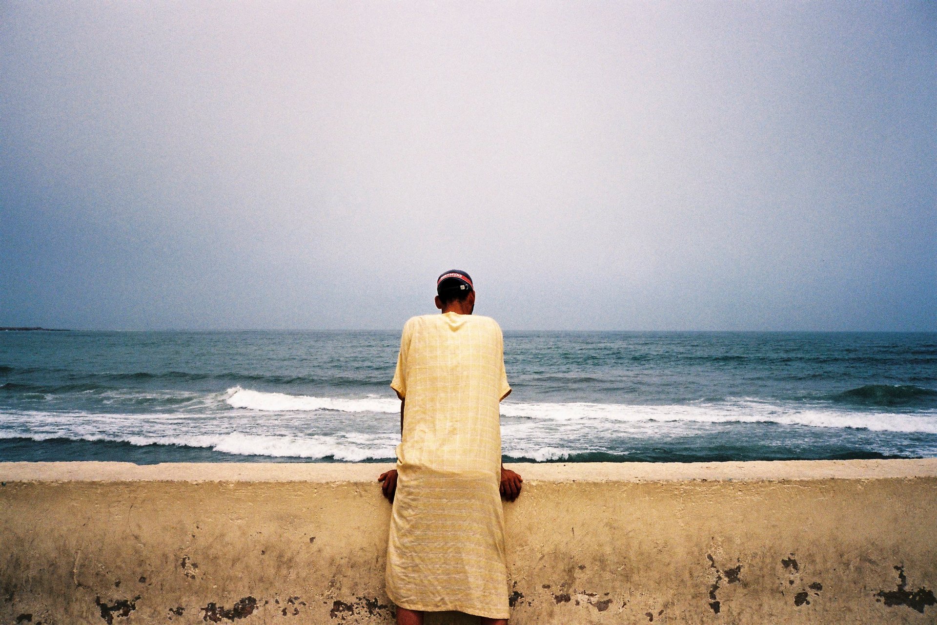 Man in traditional robe, seated on sea wall, ocean waves