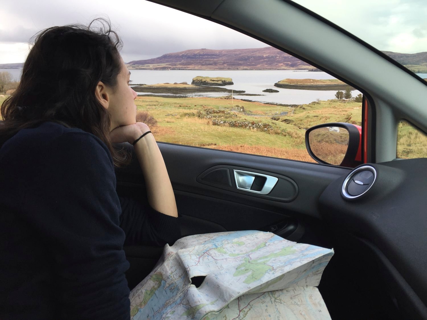 Woman in car looking out window, map on lap, loch beyond