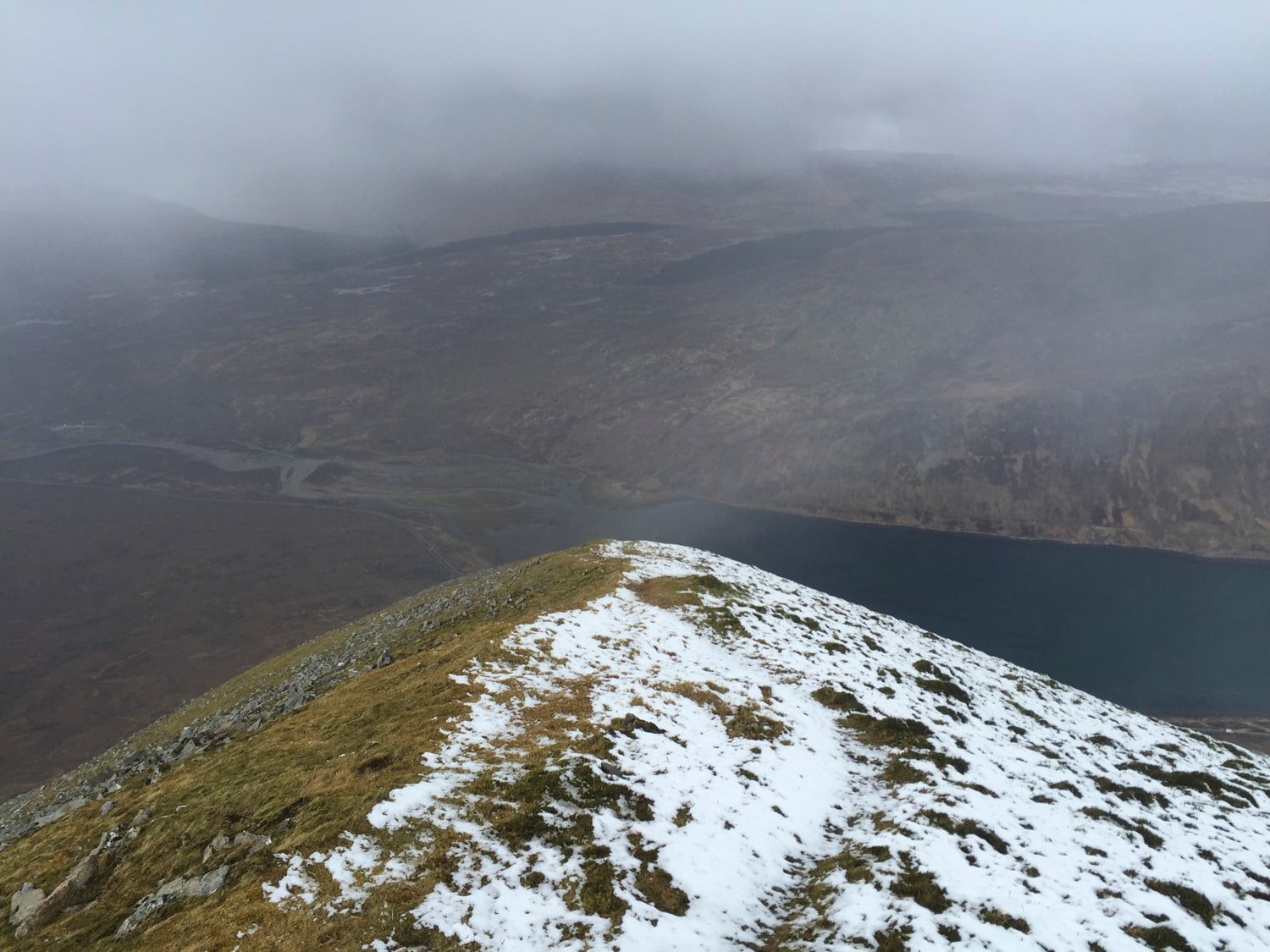 Summit view — snowy ridge, dark loch far below, mist